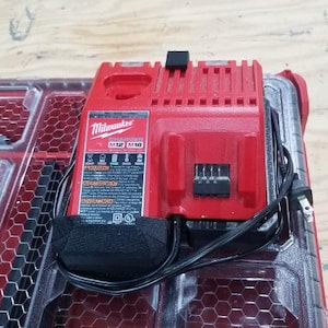 May include: A red Milwaukee M12/M18 battery charger with a black power cord and plug. The charger is on a red and clear plastic storage container. The charger has a black label with white text.