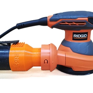 K&ouml;nnte beinhalten: Ein orangefarbener und grauer elektrischer Schleifer mit einem schwarzen Kabel und einem Staubabsauganschluss. Der Schleifer hat einen schwarzen Griff und eine graue Basis mit dem Wort "RIDGID" darauf.