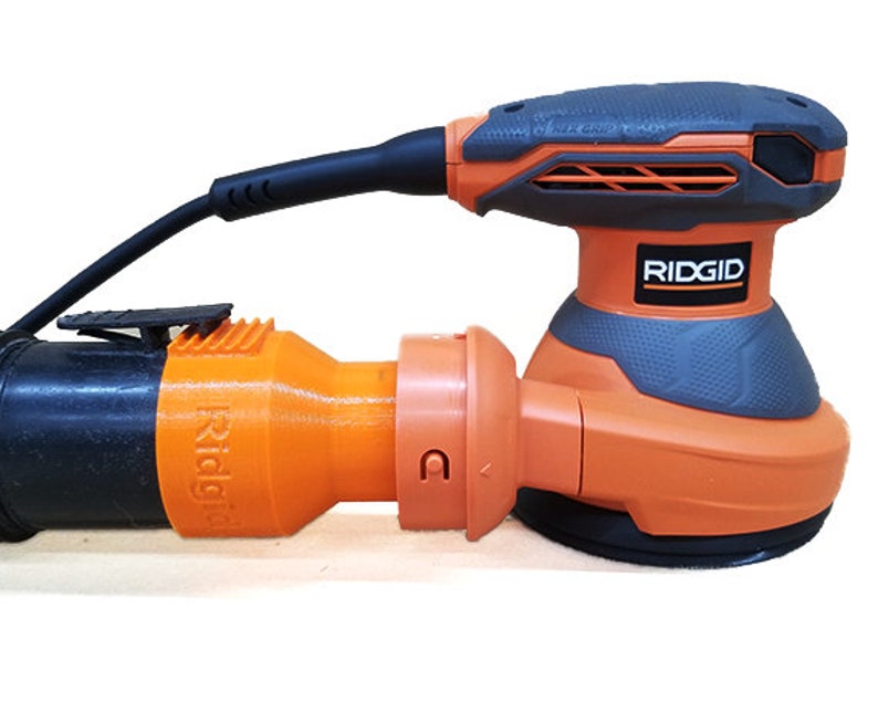 K&ouml;nnte beinhalten: Ein orangefarbener und grauer elektrischer Schleifer mit einem schwarzen Kabel und einem Staubabsauganschluss. Der Schleifer hat einen schwarzen Griff und eine graue Basis mit dem Wort "RIDGID" darauf.
