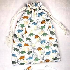 Può includere: Un sacco con coulisse bianco e cuscinetti riutilizzabili abbinati presentano un motivo di dinosauri colorati. I dinosauri sono in tonalità di verde, blu e marrone, con palme e fogliame. La borsa ha un cordoncino spesso color crema.