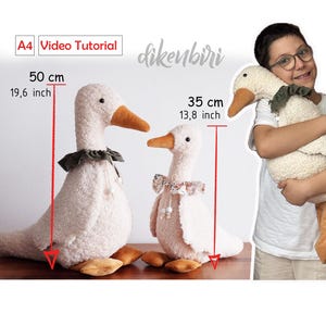 Peut inclure: Deux jouets en peluche en forme d'oie, l'un de 50 cm de haut et l'autre de 35 cm, avec des corps crème, des becs orange et des cols décoratifs. La grande oie a un col vert foncé, tandis que la petite a un col floral. L'image comprend le texte "Video Tutorial".
