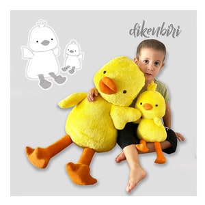 Puede incluir: Un niño sostiene un gran pato de peluche amarillo con patas y pico naranja. Un pato de peluche amarillo más pequeño con patas y pico naranja está sentado en el regazo del niño. El texto "dikenbiri" es visible en el fondo.