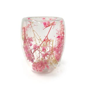 May include: A clear glass tumbler with a double-walled design, filled with pink and white dried flowers. The flowers are suspended within the glass, creating a decorative aesthetic. The tumbler is set against a white background.