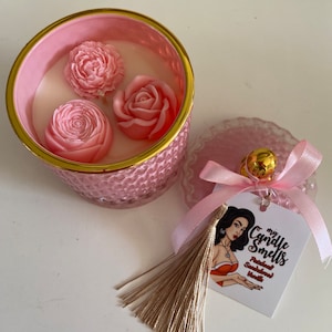 May include: Pink glass candle jar with a gold lid and three pink flower-shaped wax decorations. The jar is sitting on a white surface. A pink and gold gift tag with the text "my Candle Smells Patchouli Sandalwood Vanilla" is attached to a pink ribbon with a brown tassel.
