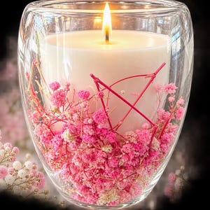 Puede incluir: Una vela encendida en un recipiente de vidrio transparente de doble pared. El espacio entre las paredes de vidrio está lleno de pequeñas flores secas de color rosa vibrante y blanco, junto a finas ramitas rojas, rodeando una cera blanca.
