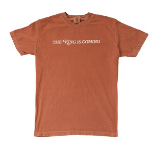 May include: A rust-colored t-shirt with the text "THE KING IS COMING" printed in white on the front.