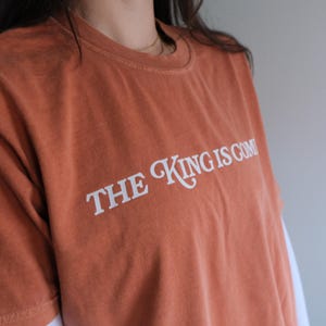 May include: A burnt orange t-shirt with the text "THE KING IS COMIN'" printed in white.