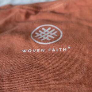 May include: A close-up of a rust-colored t-shirt with a white logo. The logo is a circle with a woven design inside. The text "WOVEN FAITH" is printed below the logo.
