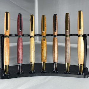 May include: Seven handcrafted wooden pens with metallic accents displayed in a black curved stand. Each pen features a unique wood grain pattern, ranging from light tan to deep purple and dark brown, paired with gold or silver clips and tips.