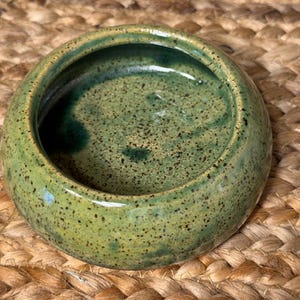 May include: A small, round ceramic bowl with a speckled green glaze. The bowl has a dark green interior and a lighter green exterior with black speckles. The bowl is sitting on a woven, tan surface.