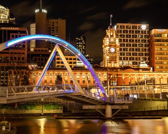 May include: A modern pedestrian bridge with blue lights arches over a river in a city at night. The bridge is lit up with blue lights and the city skyline is visible in the background. The buildings are lit up with warm yellow lights.