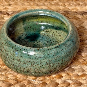 May include: A small, round ceramic bowl with a speckled, turquoise glaze. The interior has a darker, mottled green hue. The bowl is set against a woven, tan background, highlighting its handmade quality. This decorative bowl is suitable for small items.