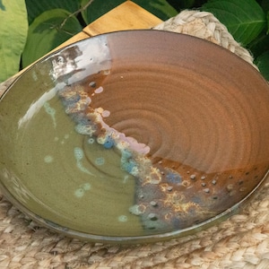 May include: A handmade ceramic plate with a two-tone design. The plate is divided into two sections: one side is a muted green, and the other is a warm brown. A band of blue, purple, and white accents separates the two colors. The plate is approximately 10 inches in diameter.