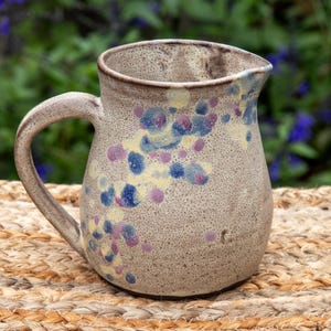 May include: A ceramic pitcher with a speckled beige base featuring a decorative pattern of blue, purple, and yellow circular spots. The pitcher has a curved handle on the side and a small pouring spout at the top rim, with a textured finish.