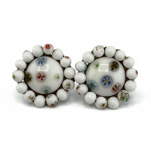 May include: A pair of vintage clip-on earrings. Each earring features a white, round center with colorful floral designs and is surrounded by a ring of white beads. The earrings have a gold-tone metal setting.