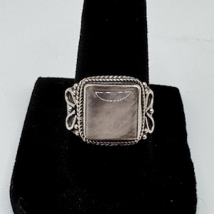 Könnte beinhalten: Ein silberner Ring mit einem quadratischen, hellfarbenen Stein. Der Ring hat einen dekorativen Rand mit geflochtenem Design und verzierten Seitendetails. Der Ring wird auf einer schwarzen Samtoberfläche präsentiert.
