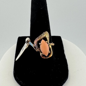 May include: A gold-toned ring featuring a marquise-shaped coral-colored stone. The ring has an open, abstract design with textured detailing on the sides. A price tag is attached.