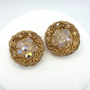 Puede incluir: Un par de pendientes vintage dorados con clip. Cada pendiente presenta un marco circular tejido que rodea un grupo de cristales claros e iridiscentes. Los pendientes tienen un diseño clásico y elegante.