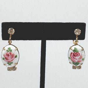 Può includere: Orecchini vintage a clip con montatura color oro. Ogni orecchino presenta un design ovale in smalto bianco con una rosa rosa e foglie verdi. Gli orecchini hanno uno strass trasparente in alto e in basso.