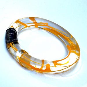 May include: Clear oval bangle bracelet with orange abstract design. The bracelet has a black label that reads "Made in England" and "Marlene Mca..." in white text. The bracelet is made of a clear material with orange streaks.