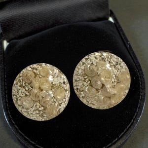 May include: A pair of round, clear resin earrings with embedded seashells and white sand-like material. The earrings are displayed in a black jewelry box with a velvet interior. The earrings have a unique, beachy aesthetic.