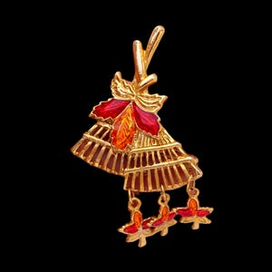May include: A gold-tone brooch featuring a fan-shaped design with red and orange enamel accents. The piece includes a central floral motif at the top and three dangling leaf-shaped charms at the bottom, all finished with a polished metallic texture.