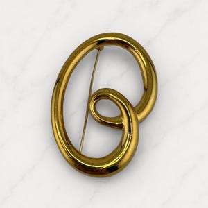May include: A gold-toned brooch with a swirling, abstract design. The brooch features a large, oval shape with a smaller, looped element and a pin closure on the back. The surface has a polished, reflective finish.
