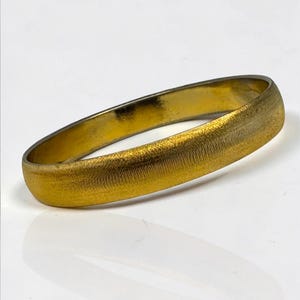May include: A gold-colored ring with a textured surface. The ring is oval-shaped and appears to be made of metal. The ring is displayed on a white surface.