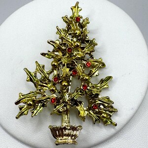 May include: A gold-toned Christmas tree brooch with red rhinestone accents. The brooch features a detailed design of a tree with holly-like leaves and small red gems. The tree sits in a decorative pot, creating a festive holiday accessory.