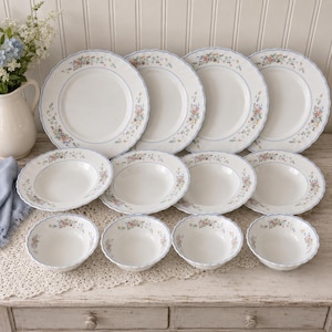 May include: A twelve-piece dinnerware set featuring four dinner plates, four soup bowls, and four small bowls. Each white ceramic piece has a scalloped edge, a thin blue rim, and a delicate floral pattern with pink, blue, and green blossoms.