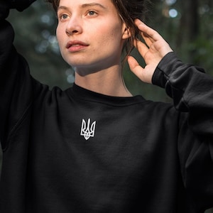 May include: A woman wearing a black sweatshirt with a white embroidered Ukrainian trident symbol. She is standing in a forest setting.