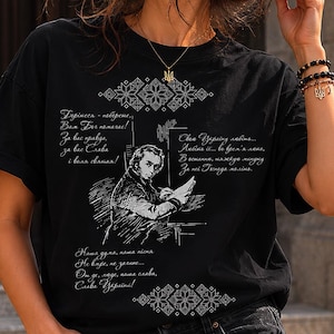 May include: Black t-shirt featuring a detailed white illustration of a man writing, with text in a foreign language. The shirt also has decorative patterns and additional text. The shirt is worn by a person.