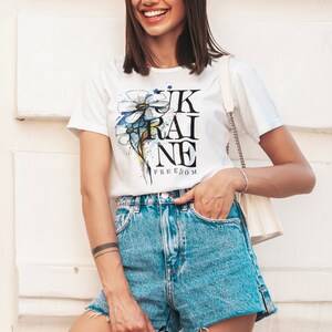 Peut inclure: Une femme portant un t-shirt blanc avec un motif de fleur bleu et jaune et le texte "UKRAINE FREEDOM". Elle porte &eacute;galement un short en jean et tient des lunettes de soleil.