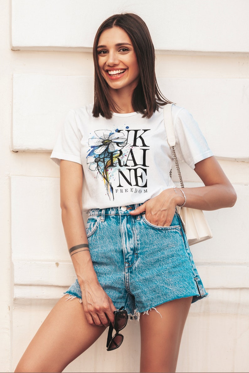Peut inclure: Une femme portant un t-shirt blanc avec un motif de fleur bleu et jaune et le texte "UKRAINE FREEDOM". Elle porte &eacute;galement un short en jean et tient des lunettes de soleil.