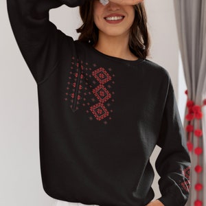 May include: Black sweatshirt with a red embroidered design on the front. The design features geometric shapes and floral patterns.