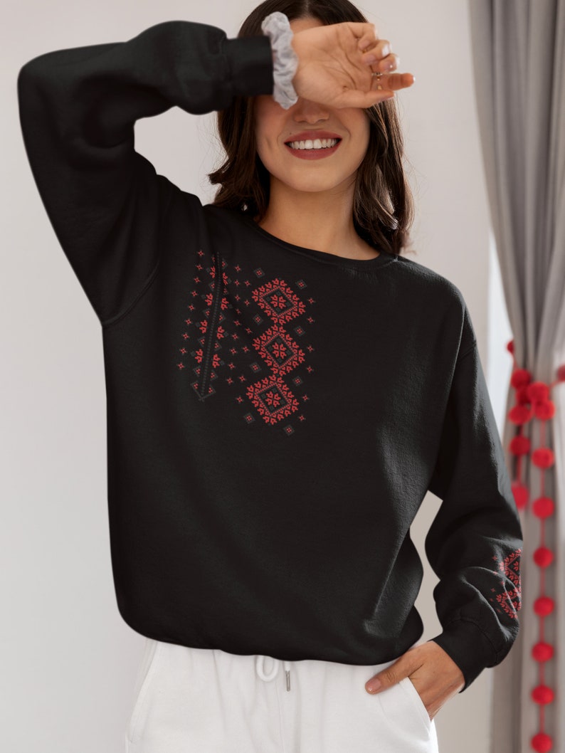 May include: Black sweatshirt with a red embroidered design on the front. The design features geometric shapes and floral patterns.