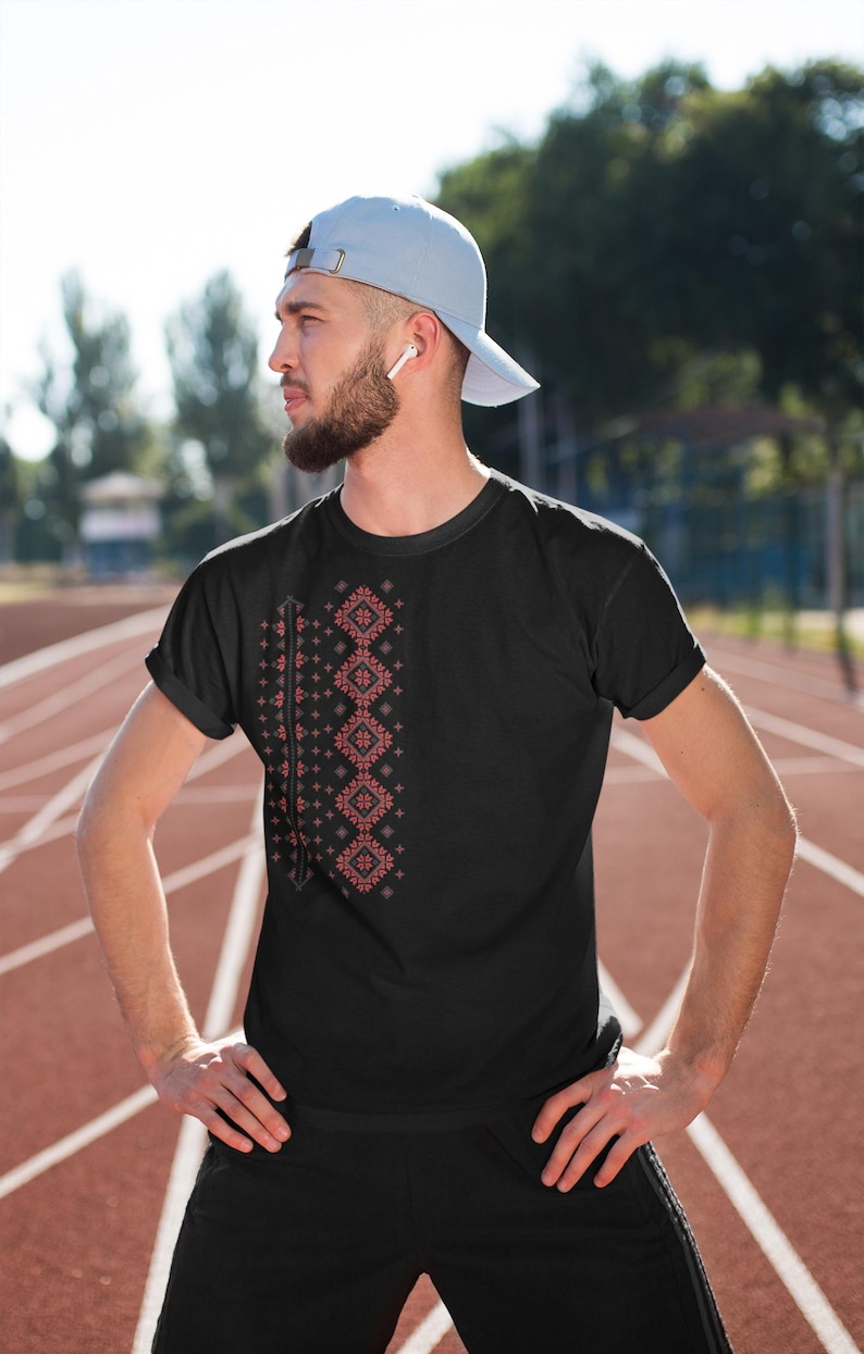 Puede incluir: Camiseta negra con un dise&ntilde;o geom&eacute;trico rojo y blanco en el lado izquierdo. La persona lleva una gorra de b&eacute;isbol azul claro y pantalones deportivos negros. El fondo es una pista de atletismo roja.