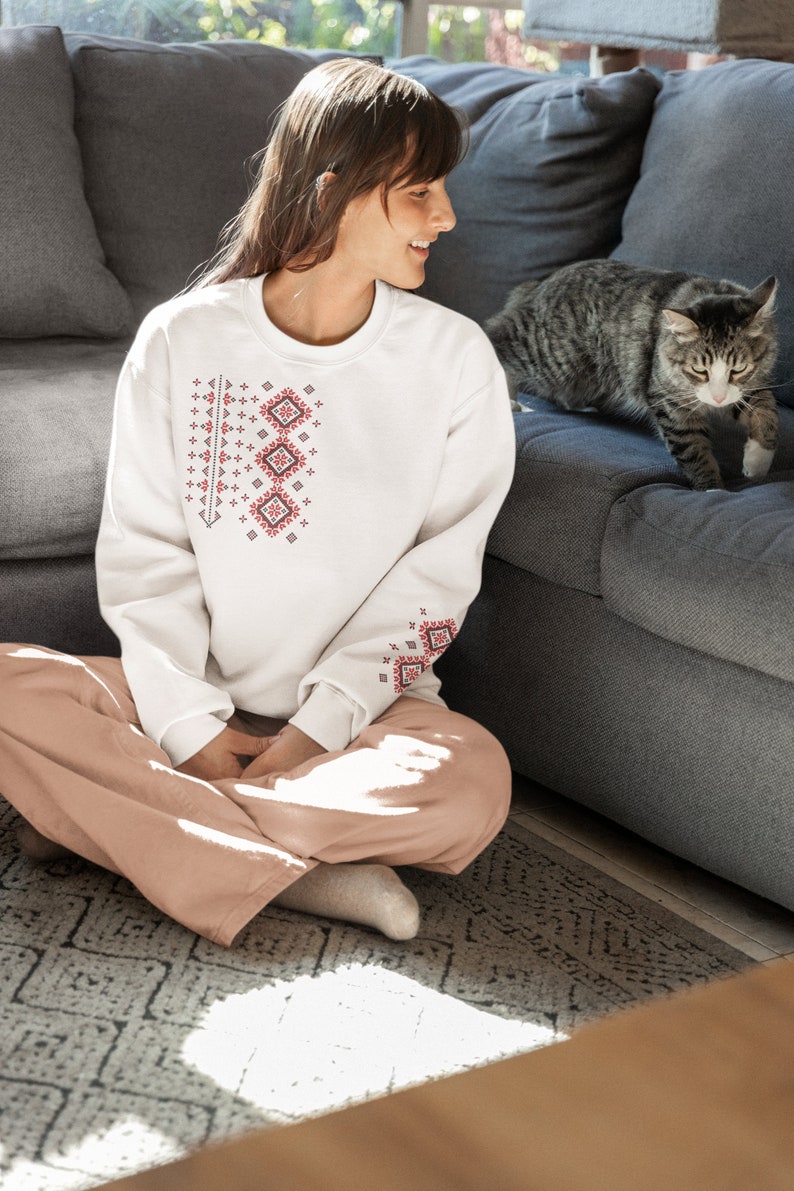 May include: A woman wearing a white sweatshirt with a red and white embroidered design sits on a gray couch with a gray tabby cat. The woman is wearing pink pants and is sitting cross-legged. The cat is sitting on the couch and looking at the woman.