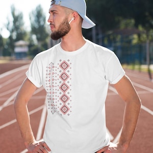 Puede incluir: Camiseta blanca con un dise&ntilde;o bordado geom&eacute;trico rojo y negro en el lado izquierdo. El dise&ntilde;o presenta patrones de diamantes y cruces. La camiseta la lleva una persona que lleva una gorra de b&eacute;isbol azul claro.