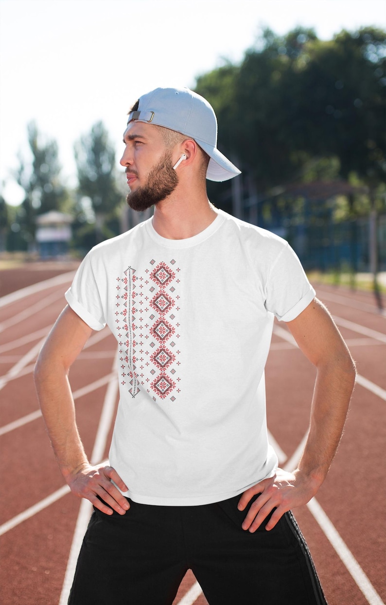 Puede incluir: Camiseta blanca con un dise&ntilde;o bordado geom&eacute;trico rojo y negro en el lado izquierdo. El dise&ntilde;o presenta patrones de diamantes y cruces. La camiseta la lleva una persona que lleva una gorra de b&eacute;isbol azul claro.