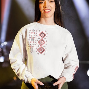 May include: A woman wearing a white embroidered shirt with red and black designs holds a black book with the text "Santa Biblia" on the cover.