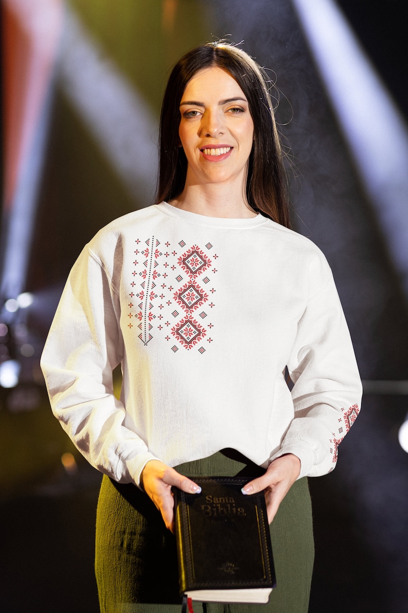 May include: A woman wearing a white embroidered shirt with red and black designs holds a black book with the text "Santa Biblia" on the cover.