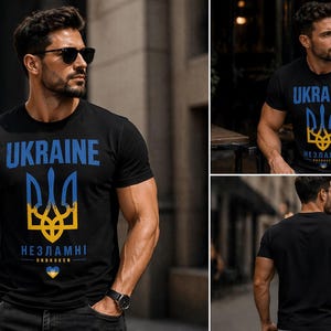Op de afbeelding: Zwart T-shirt met het woord "UKRAINE" in blauw, boven een blauw en geel drietandsymbool. Daaronder staat het woord "НЕЗЛАМНІ" in wit, met een kleine hartafbeelding. Het T-shirt wordt door een persoon gedragen.