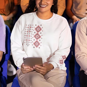 May include: A woman wearing a white sweatshirt with a red and black geometric pattern sits in a blue chair and holds a tablet.