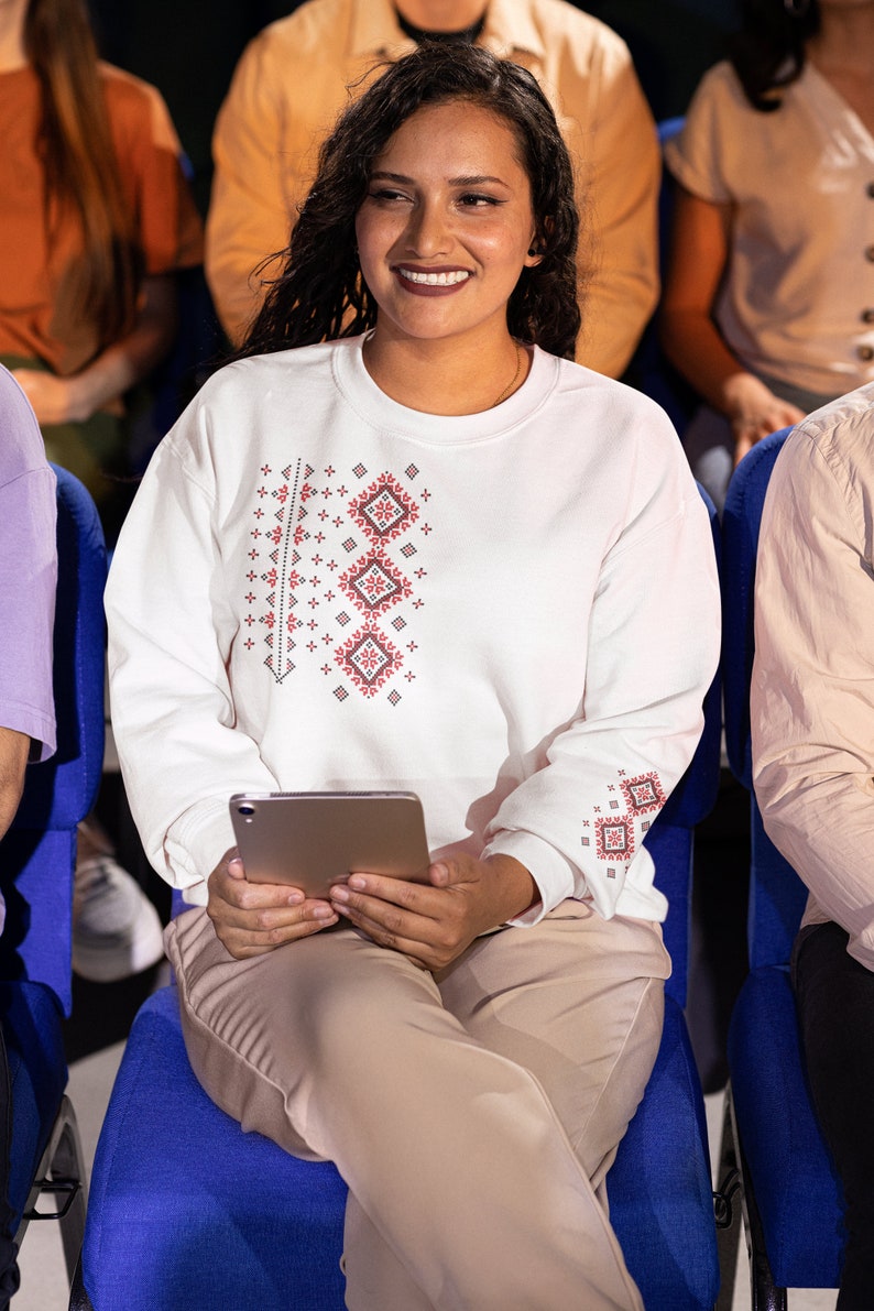 May include: A woman wearing a white sweatshirt with a red and black geometric pattern sits in a blue chair and holds a tablet.