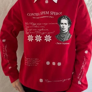May include: Red sweatshirt with a white collar, adorned with a portrait of a woman and white text. The text includes "CONTRA SPEM SPERO!" and other phrases in a script. White star and geometric designs are also featured.