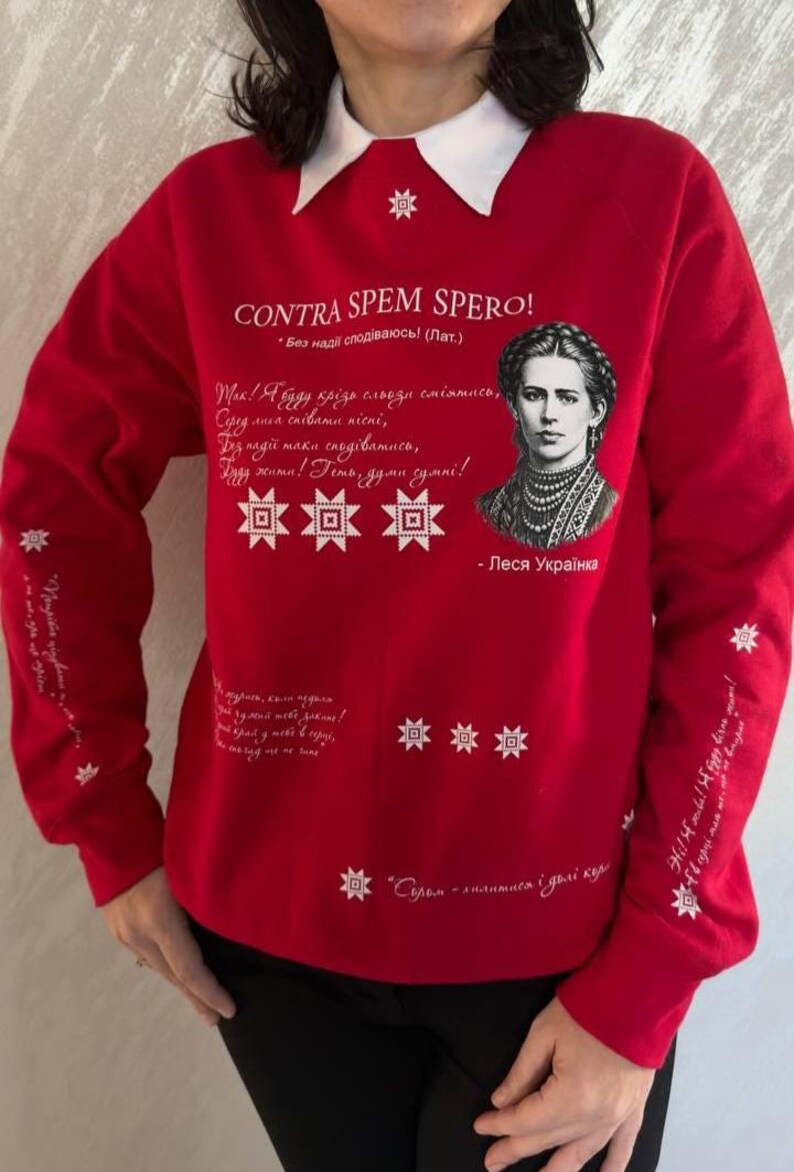 May include: Red sweatshirt with a white collar, adorned with a portrait of a woman and white text. The text includes "CONTRA SPEM SPERO!" and other phrases in a script. White star and geometric designs are also featured.