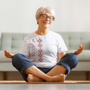 Puede incluir: Una camiseta blanca con un dise&ntilde;o geom&eacute;trico rojo y gris, combinada con pantalones azules. La persona est&aacute; sentada con las piernas cruzadas sobre una esterilla, con los ojos cerrados y las manos en posici&oacute;n de meditaci&oacute;n. El fondo incluye un sof&aacute; claro.