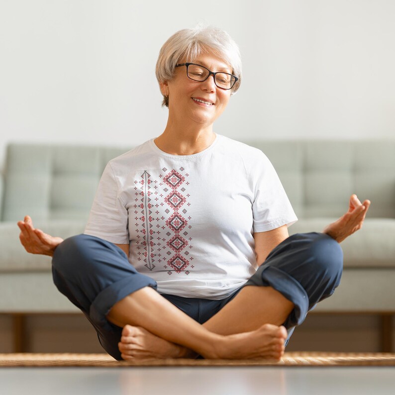 Puede incluir: Una camiseta blanca con un dise&ntilde;o geom&eacute;trico rojo y gris, combinada con pantalones azules. La persona est&aacute; sentada con las piernas cruzadas sobre una esterilla, con los ojos cerrados y las manos en posici&oacute;n de meditaci&oacute;n. El fondo incluye un sof&aacute; claro.