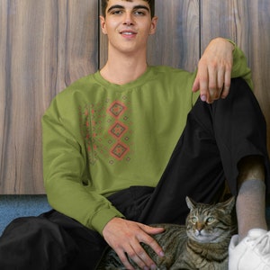 May include: A man wearing a green sweatshirt with a red and black geometric pattern sits on the floor with a tabby cat. He is wearing black pants and white sneakers.